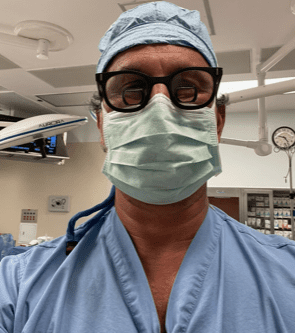 A man in scrubs and mask standing inside of an operating room.